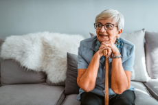 "Gesund im Pflegeheim" Foto einer alten Frau mit grauen Haaren, die mit ihrem Gehstock auf einem Sofa sitzt und weg schaut. 