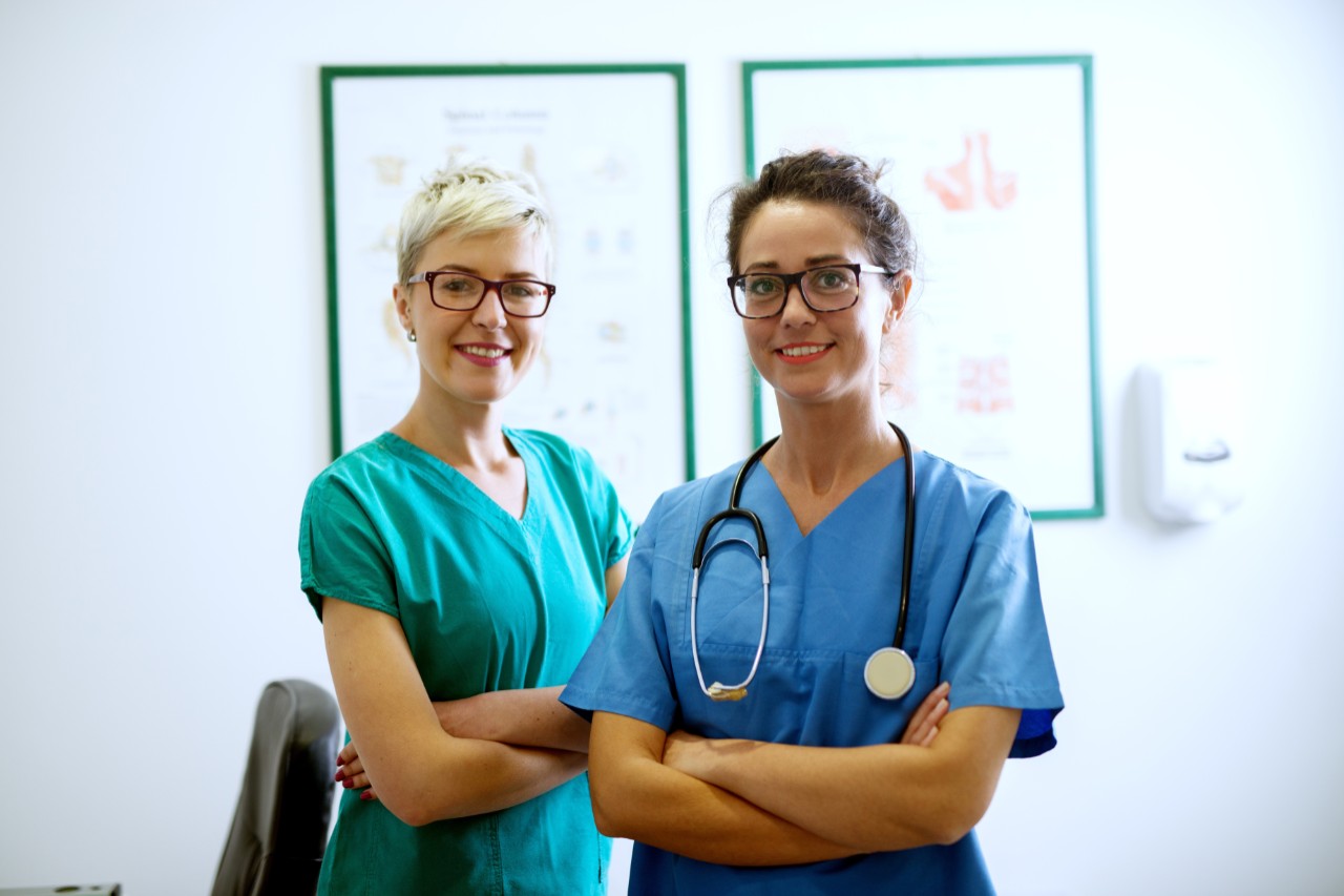 Portrait zweier fröhlicher Krankenschwestern mit Brillen, die mit verschränkten Armen im Büro vor zwei Bilderrahmen stehen und in die Kamera schauen. Portrait zweier fröhlicher Krankenschwestern mit Brillen, die mit verschränkten Armen im Büro vor zwei Bilderrahmen stehen und in die Kamera schauen.