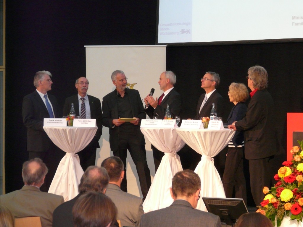 Blick aufs Podium mit den Diskussionsteilnehmern, Landrat Thomas Reumann hält ein Mikrofon und spricht Blick aufs Podium mit den Diskussionsteilnehmern, Landrat Thomas Reumann hält ein Mikrofon und spricht