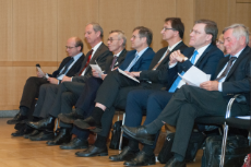 Sieben Männer in Anzügen sitzen in einer Stuhlreihe und hören zu