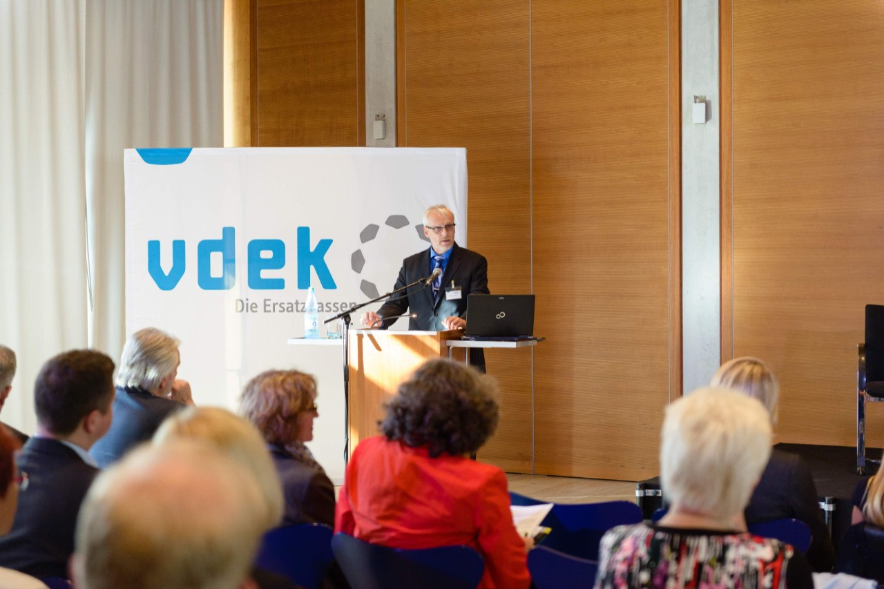 Blick aus dem Publikum aufs Podium, Michael Domr&ouml;s steht am Rednerpult und spricht