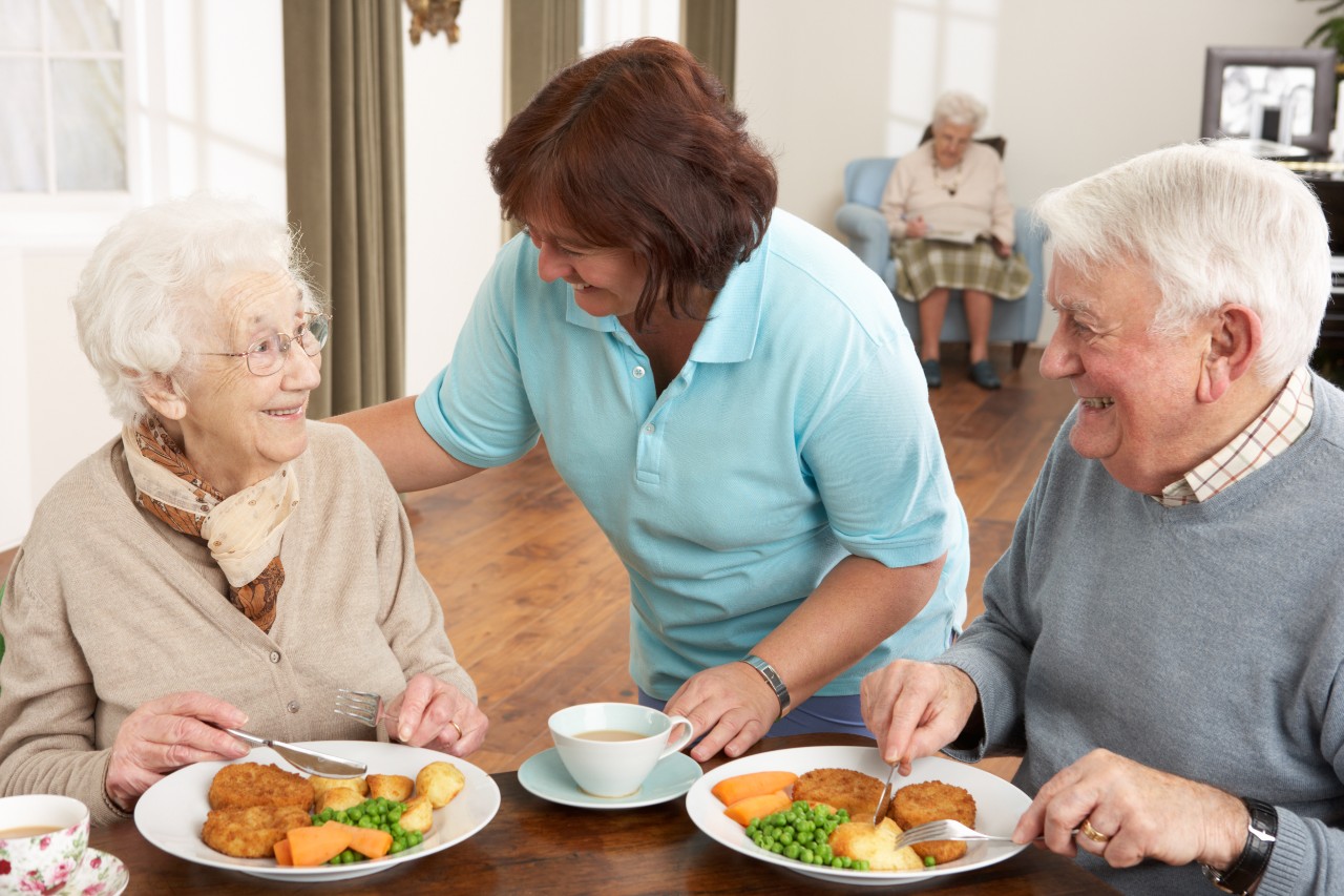 Senioren beim Mittagessen; Pflegekraft hilft