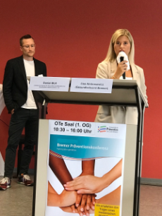Frau mit Mikro am Stehtisch und Mann im Hintergrund