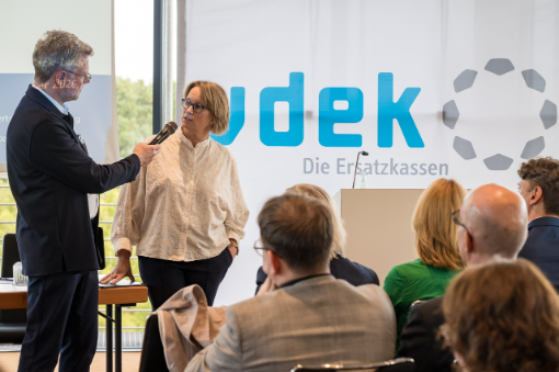 Frau wird von Mann mit Mikrofon interviewt vor Publikum in Saal
