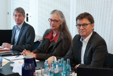 Zwei Männer und eine Frau sitzen an einem Tisch und hören zu