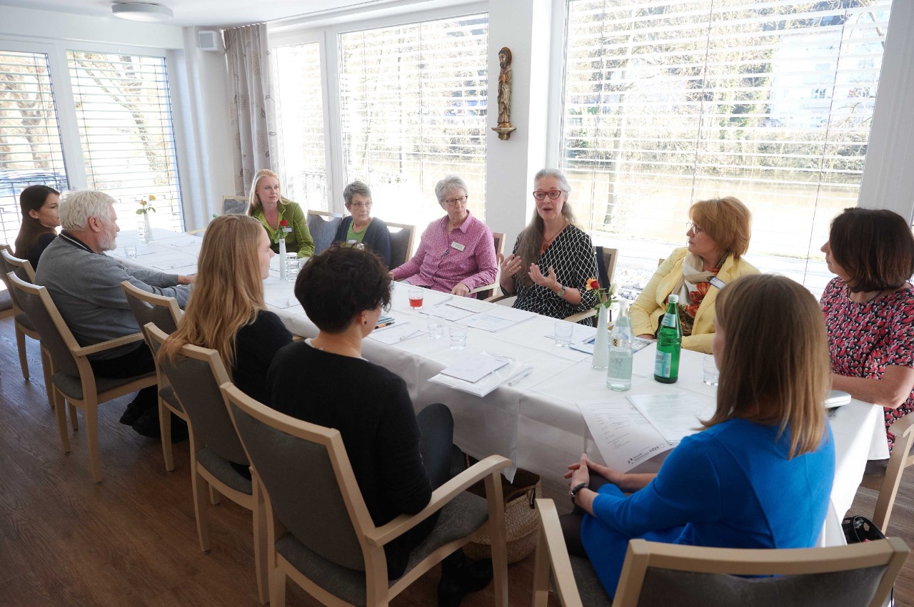 An einem langen Tisch sitzen zu beiden Seiten sechs Personen. Bis auf eine Person sind alles Frauen. An einem langen Tisch sitzen zu beiden Seiten sechs Personen. Bis auf eine Person sind alles Frauen.