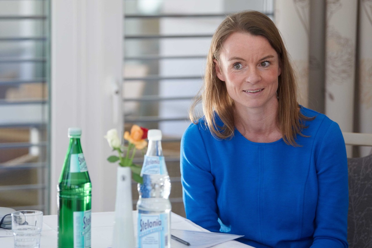 Eine Frau mit blauem Kleid und langen hellbraunen Haaren sitzt an einem Tisch und redet. Eine Frau mit blauem Kleid und langen hellbraunen Haaren sitzt an einem Tisch und redet.