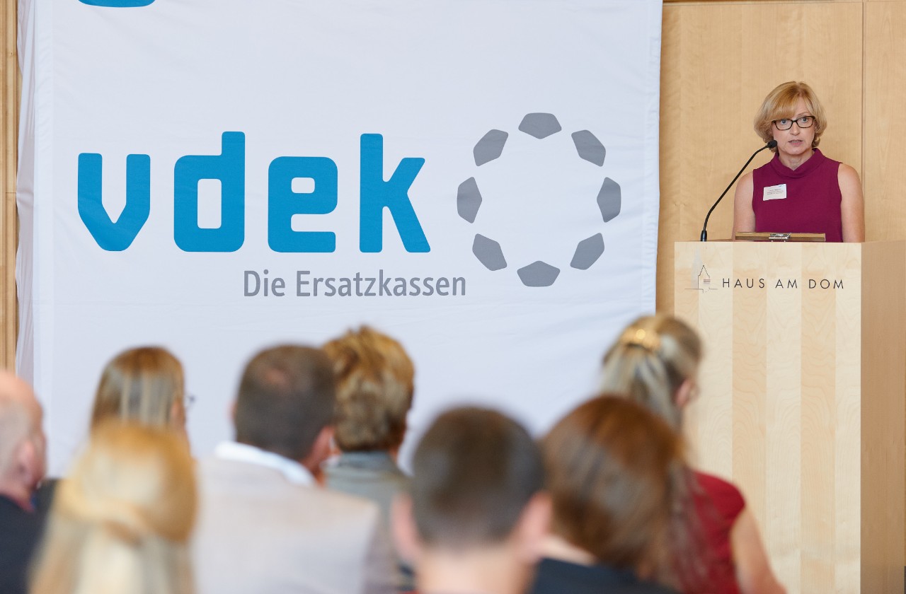 Frau mit kurzen Haaren steht hinter einem Stehpult und spricht zu sitzenden Menschen Frau mit kurzen Haaren steht hinter einem Stehpult und spricht zu sitzenden Menschen