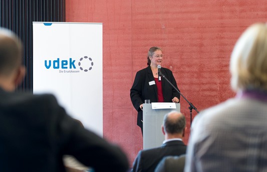 Blick zwischen den K&ouml;pfen des Publikums hindurch auf das Rednerpult, an welchem Claudia Ackermann steht und spricht