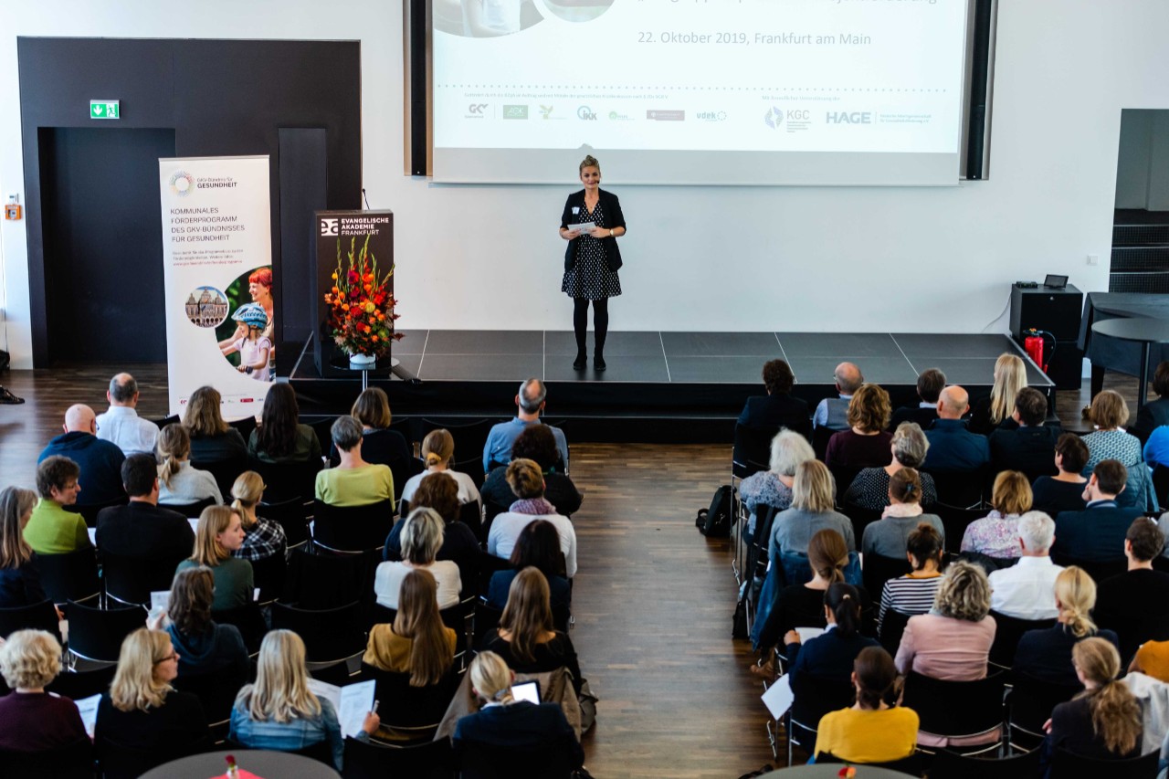 blonde Frau auf Bühne spricht zu Menschen, die auf Stühlen in einem vollen Saal sitzen