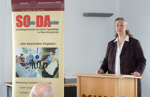 Claudia Ackermann steht am Rednerpult und spricht, links von ihr steht ein Banner des Projektes &quot;SOwieDAheim&quot;