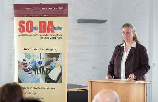 Claudia Ackermann steht am Rednerpult und spricht, links von ihr steht ein Banner des Projektes &quot;SOwieDAheim&quot;