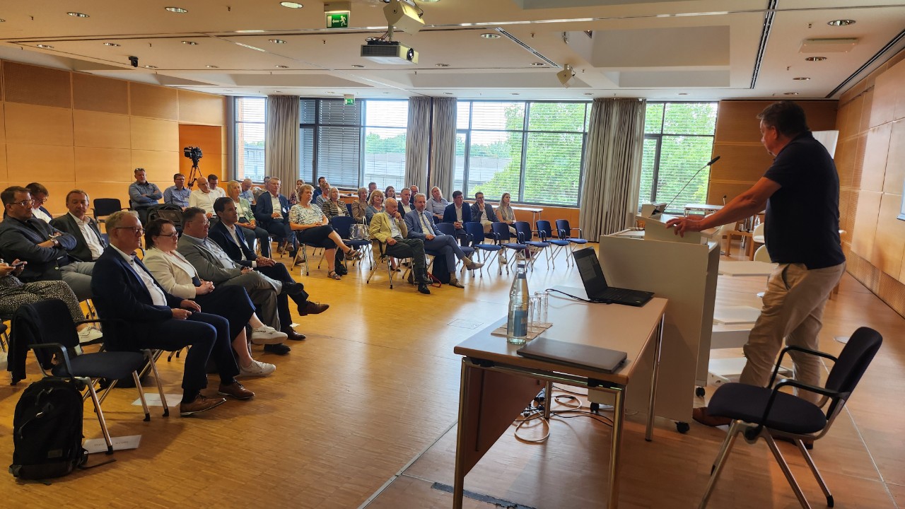 Zuschauende auf St&uuml;hlen h&ouml;ren einem Redner am Pult zu