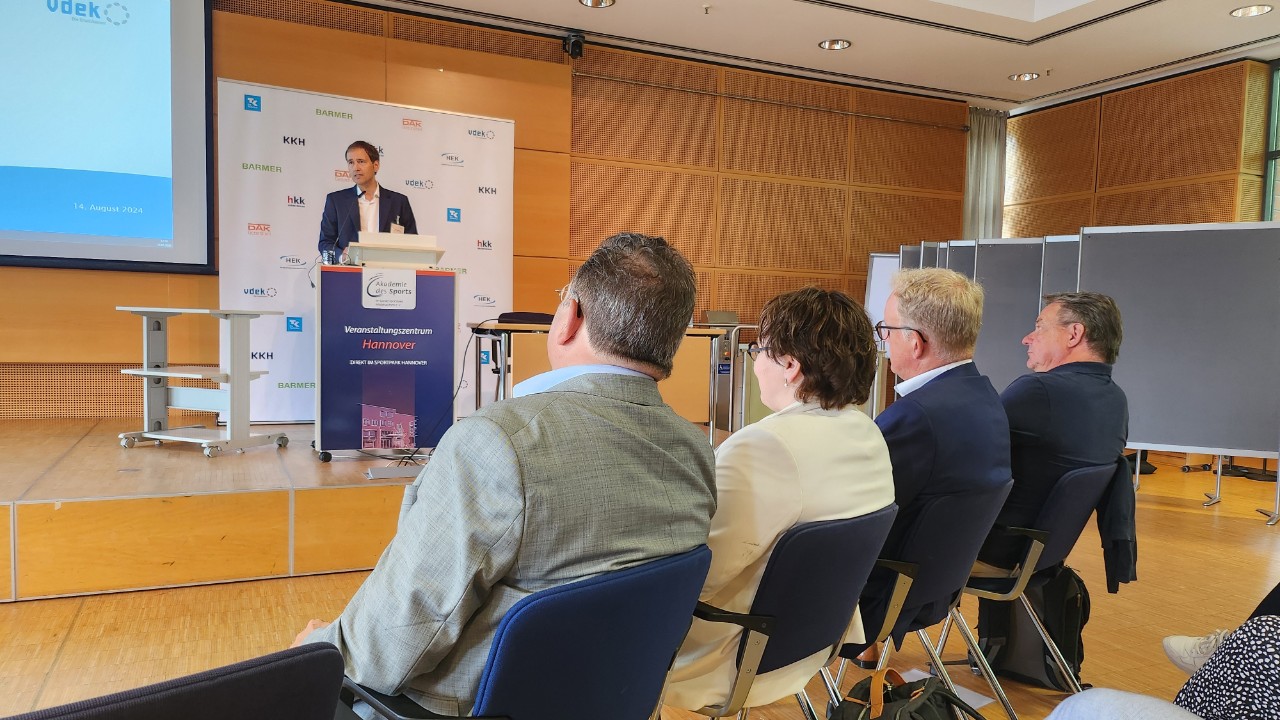 Vier Personen sitzen auf St&uuml;hlen und h&ouml;ren einem Redner zu