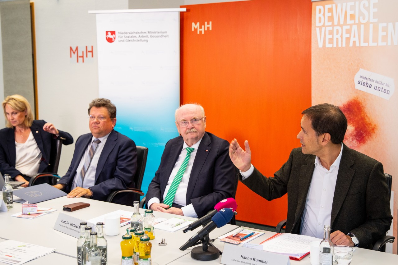 Drei Männer und eine Frau Sitzen an einem Tisch mit Mikrofonen und Dokumenten