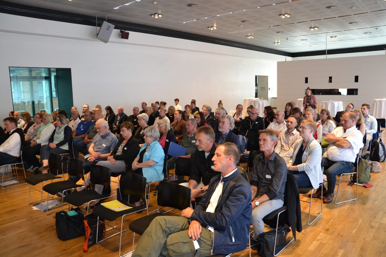 Viele Zuschauende sitzen auf St&uuml;hlen in mehreren Reihen und gucken nach vorne.