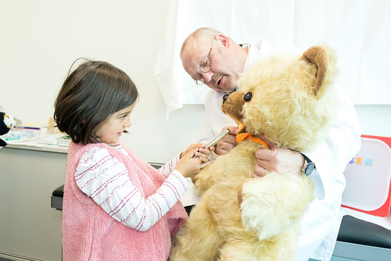 Arzt hat einen großen Teddy auf dem Schoss und spricht mit kleinem Mädchen