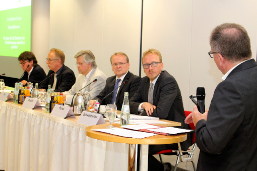 Fünf Männer sitzen an einem Podiumstisch, ein Mann steht mit Mikro rechts daneben