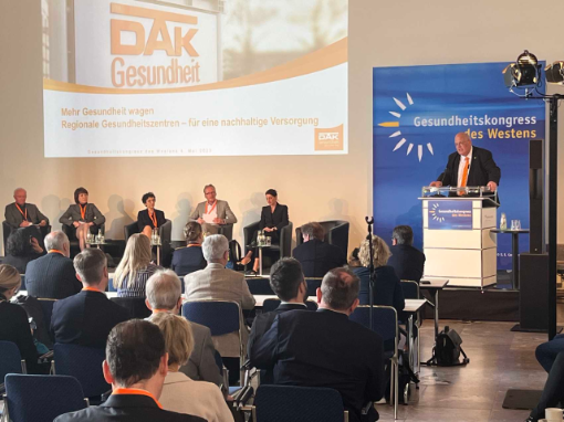 Fünf Personen auf dem Podium, ein Mann am Rednerpult, im Vordergrund sitzendes Publikum