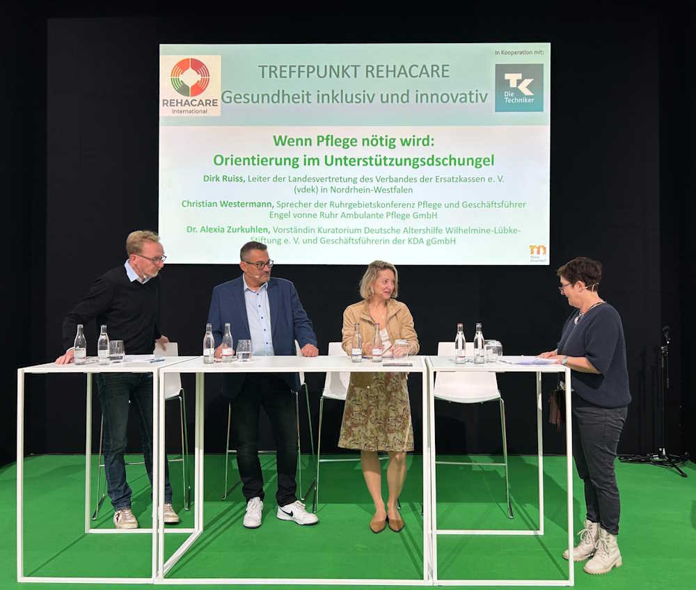 Drei Menschen hinter einem Stehtisch auf einem Podium, rechts daneben eine Moderatorin