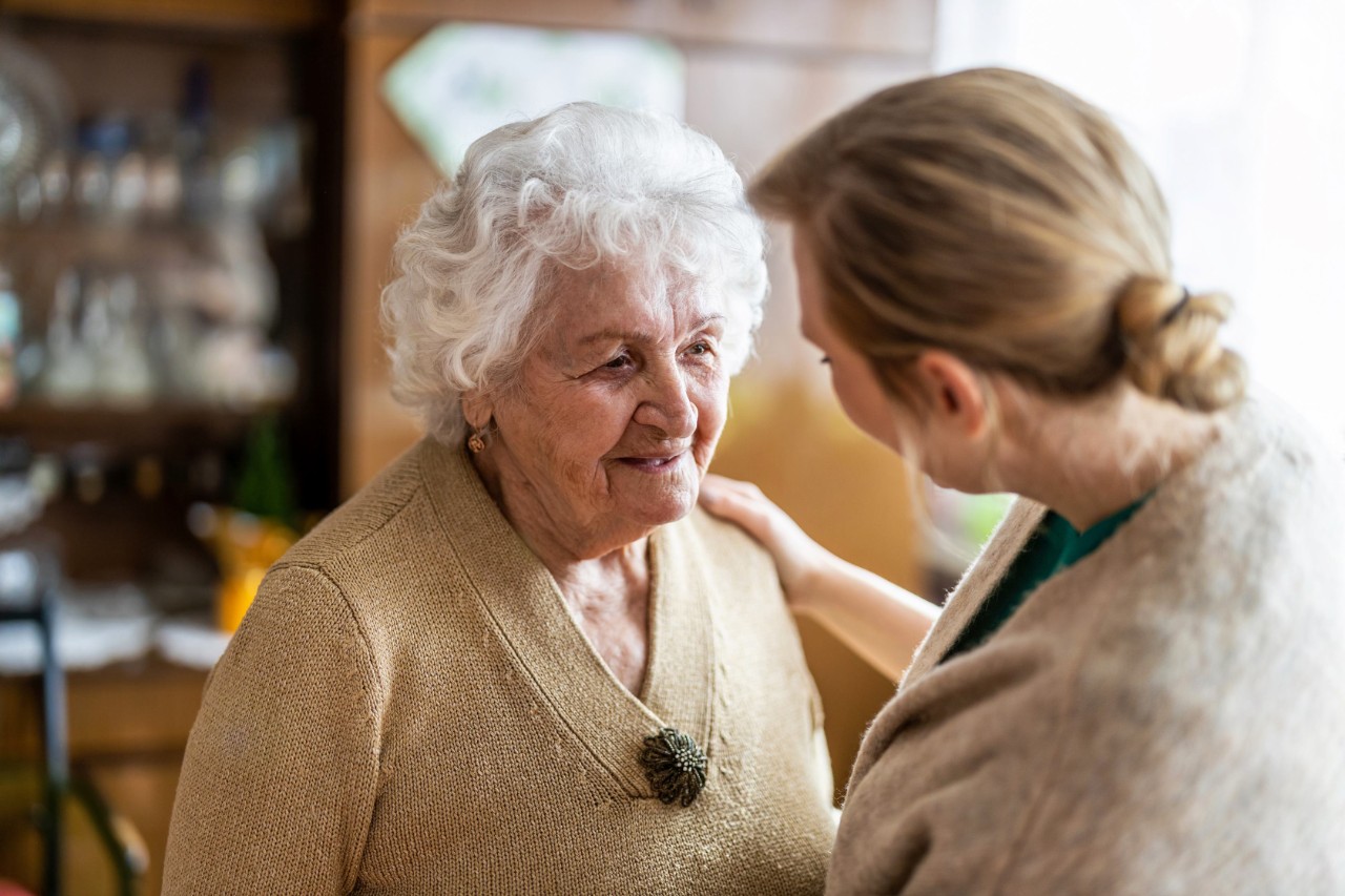 Jüngere Frau im Gespräch mit einer älteren Frau. Im Hintergrund ist ein Rollator erkennbar.