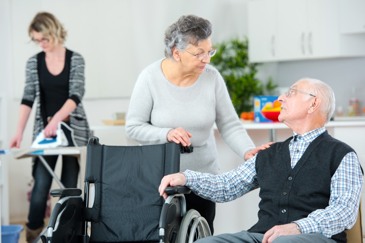 Häusliche Pflege, Im Vordergrund sitzt ein alter Mann neben einem Rollstuhl, hinter dem eine alte Frau steht. Die beiden schauen sich an. Im Hintergrund bügelt eine junge Frau mit Bügeleisen auf einem Bügelbrett vor einer Küchenzeile.