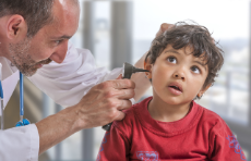 Beim Kinderarzt, Arzt im Arztkittel untersucht mit Untersuchungsinstrument Ohr von kleinem Jungen in rotem Pullover