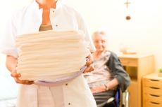 Pflegeheim, Junge Pflegerin steht im Vordergrund mit Handtüchern, im Hintergrund sitzt ältere Dame im Rollstuhl