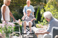 Senioren treffen sich im Garten, Pflegerin schiebt Frau im Rollstuhl, Mann sitzt auf Parkbank, Frau steht mit Gehhilfe