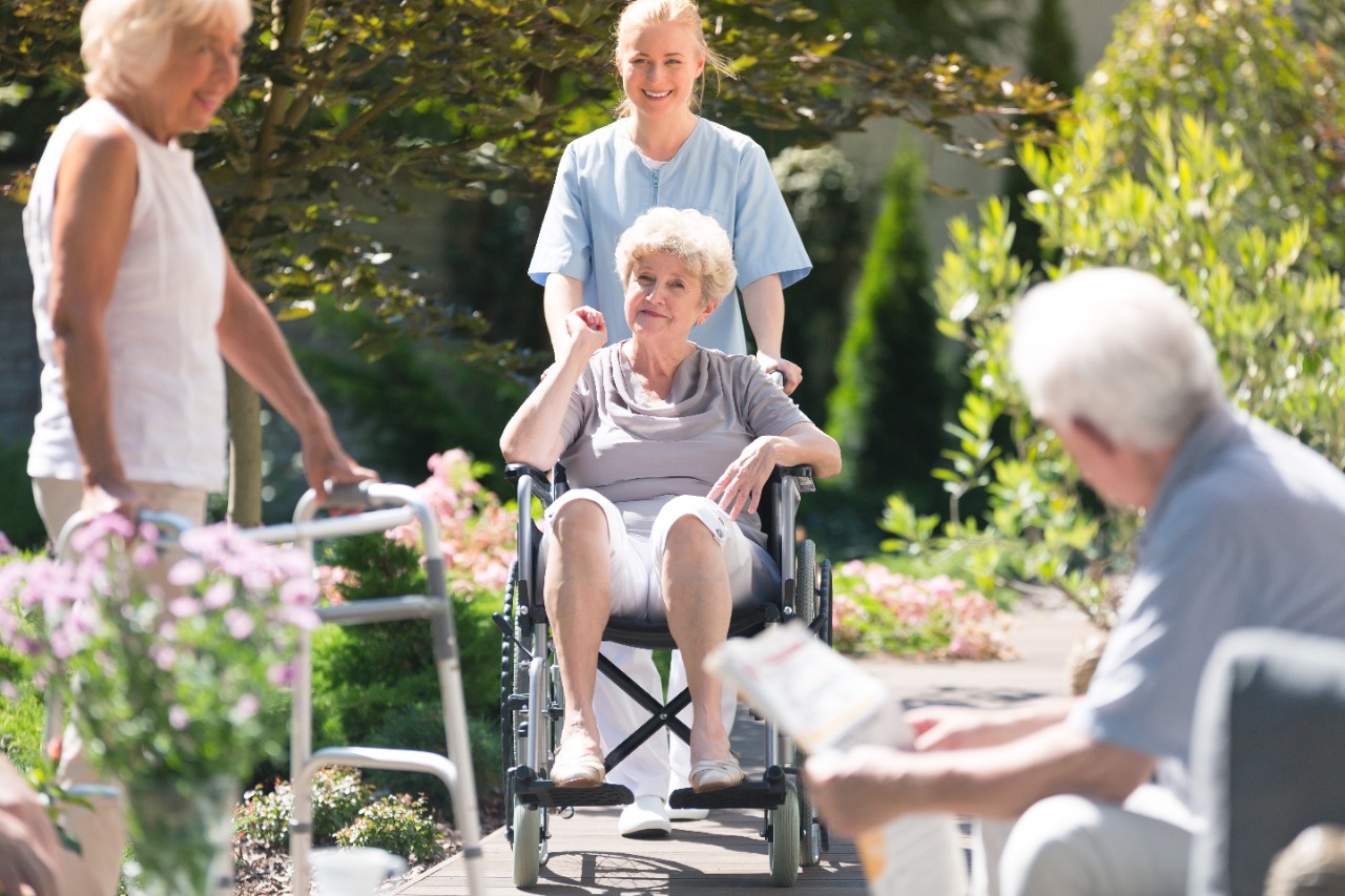 Senioren treffen sich im Garten, Pflegerin schiebt Frau im Rollstuhl, Mann sitzt auf Parkbank, Frau steht mit Gehhilfe Senioren treffen sich im Garten, Pflegerin schiebt Frau im Rollstuhl, Mann sitzt auf Parkbank, Frau steht mit Gehhilfe