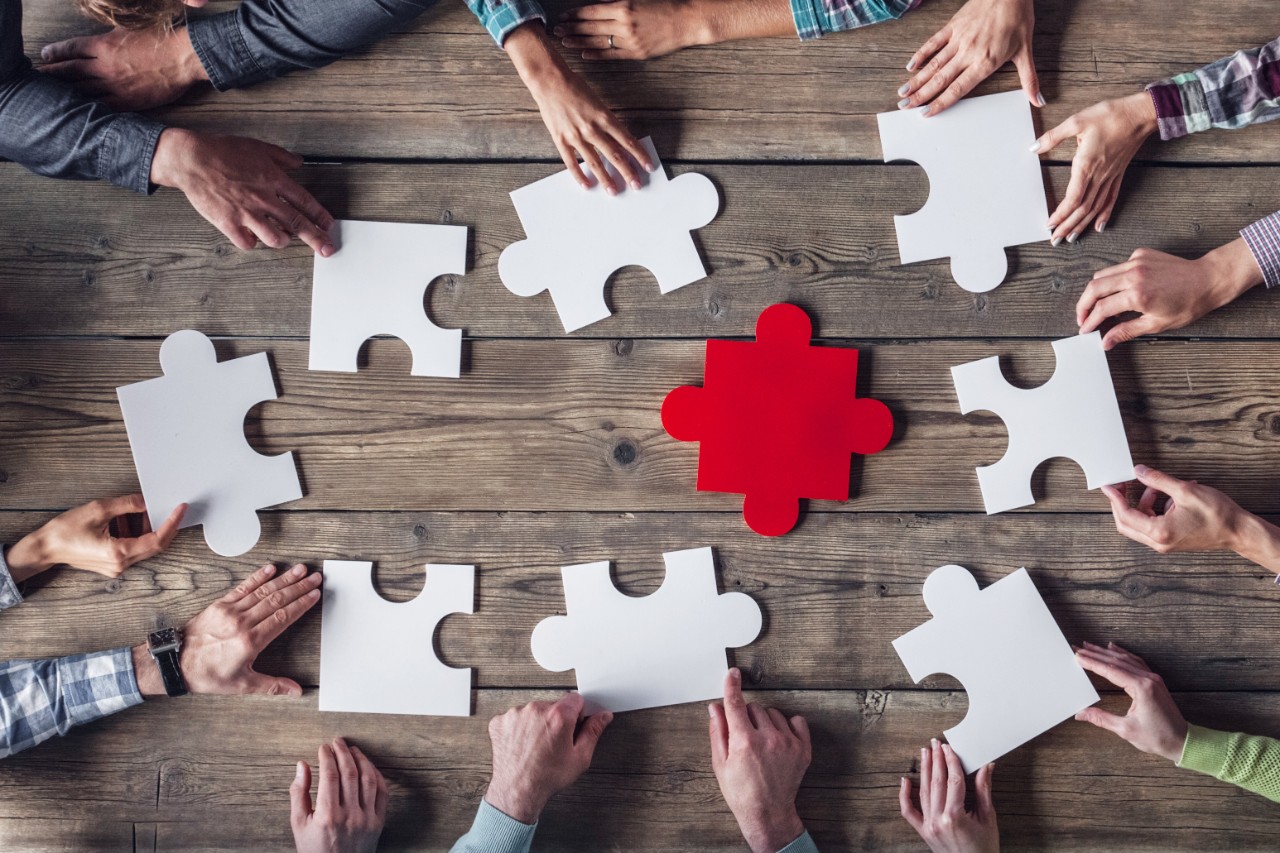 Teamwork Meeting, Die Hände von acht im Kreis an einem Holztisch sitzenden Personen halten jeweils ein großes weißes Puzzlestück. In der Mitte des Tisches liegt ein großes rotes Puzzleteil. Teamwork Meeting, Die Hände von acht im Kreis an einem Holztisch sitzenden Personen halten jeweils ein großes weißes Puzzlestück. In der Mitte des Tisches liegt ein großes rotes Puzzleteil.
