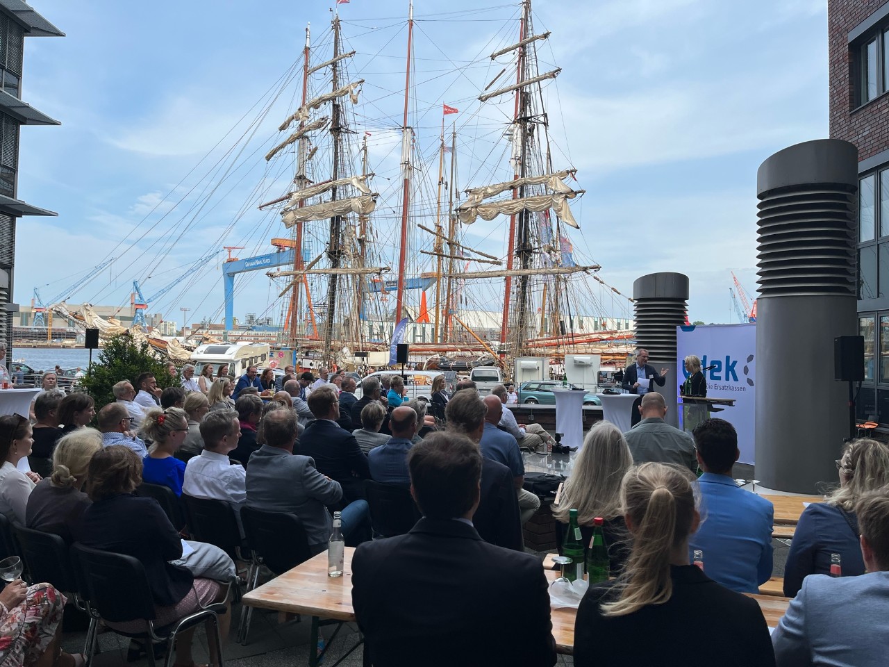 Viele Menschen sitzen auf Stühlen und Bänken in einem Innenhof unter freiem Himmel - im Hintergrund liegt ein Segelschiff