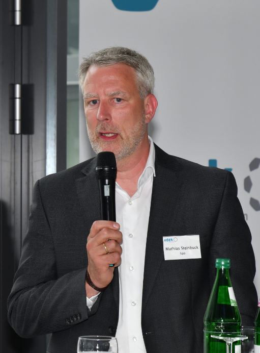 Ein Mann im weißen Oberhemd und dunklen Anzug spricht in ein Mikrofon, das er in der Hand hält.