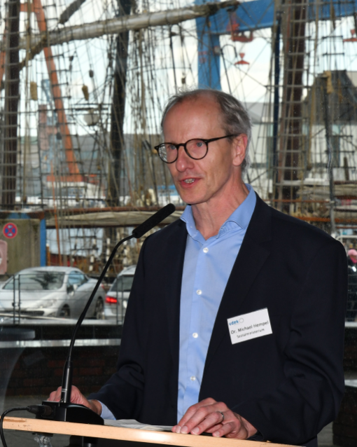 Ein Mann mit Brille im dunklen Anzug und hellblauem Oberhemd steht an einem Rednerpult. Im Hintergrund sieht man die Takelage eines Segelschiffes und den Portalkran der Kieler Werft.