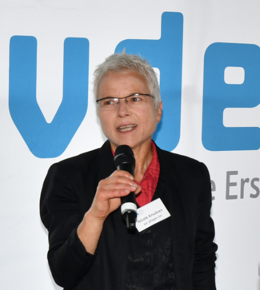 Eine Frau in einem schwarzen Blazer steht vor dem Logo des vdek und spricht in ein Mikrofon, das sie in der Hand hält.