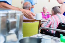 Hand eines Pflegers, die eine Tasse hält. Im Hintergrund sitzen Pflegebedürftige an einem Tisch