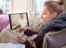 Junge Frau liegt krank auf dem Sofa mit einem Tablet und hat eine Videosprechstunde mit einem Arzt