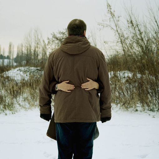 Ein Mann steht mit dem Rücken zur Kamera in Winterlandschaft. Auf seinem Rücken zwei Hände von einer Frau, die ihn umarmt.