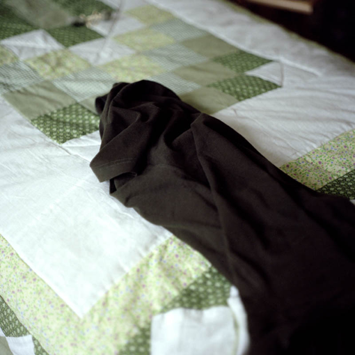 Ausschnitthafter Blick auf ein gemachtes Bett, am Rand liegt ein Kleidungsstück.