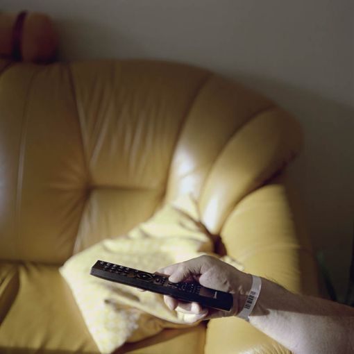 Wohnzimmersofa im Hintergrund, im Vordergrund ein ausgestreckter Arm mit einer Fernsehbedienung