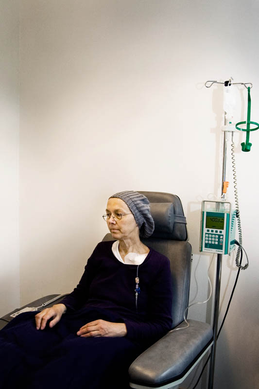 Eine Frau sitzt auf einem Sessel. Daneben steht ein Infusionsgerät, der Kopf ist mit einer Mütze bedeckt.