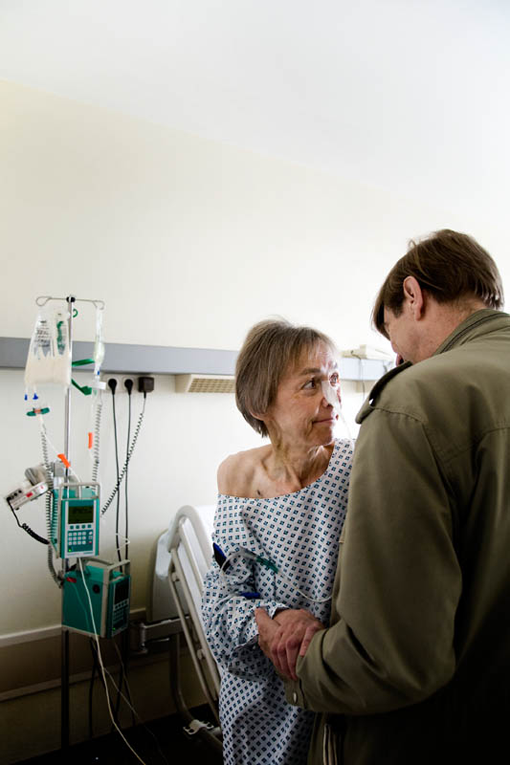 Eine Frau steht neben einem Krankenhausbett einem Mann zugewandt. Sie halten die Hände, im Hintergrund ein Infusionsgerät.