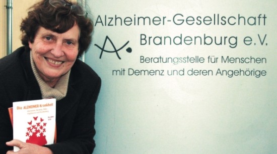 Eine Frau steht vor einem Schild mit der Aufschrift "Alzheimer-Gesellschaft Brandenburg e. V. Beratungsstelle für Menschen mit Demenz und deren Angehörige"