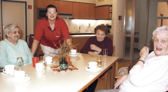 Drei alte Damen sitzen um einen Küchentisch, eine Betreuerin steht dabei