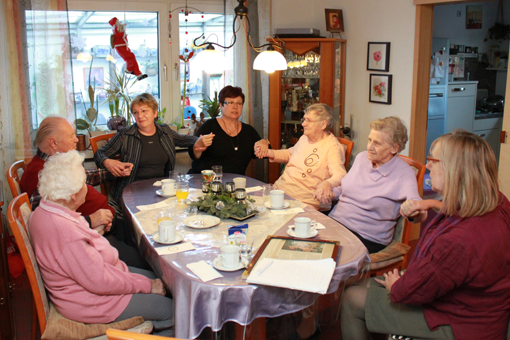 Eine Gruppe älterer Frauen sitzen um einen Wohnzimmertisch und halten sich die Hände