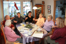 Eine Gruppe älterer Frauen sitzen um einen Wohnzimmertisch und halten sich die Hände