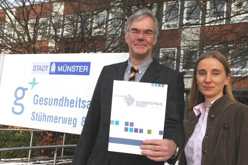 Frau und Mann stehen vor einem Haus und einem Schild mit der Aufschrift &quot;Stadt M&uuml;nster Gesundheitsamt&quot;. Der Mann h&auml;lt eine Publikation in die Kamera.