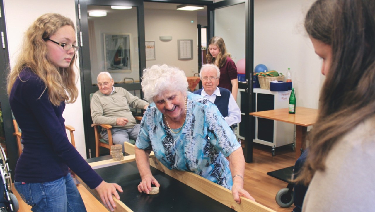 Szene aus dem Seniorenheim, zwei junge Mädchen spielen mit älteren Frauen und Männern