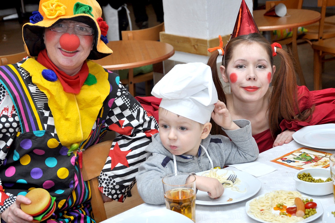 Zwei Frauen Erzieherinnen im Clowns-Kost&uuml;m knien hinter einem Kind mit Kochm&uuml;tze am Mittagstisch