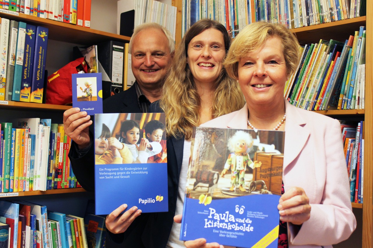 Vor einem B&uuml;cherregal halten zwei Frauen und ein Mann jeweils ein Buch &uuml;ber das Pr&auml;ventionsprogramm Papilio in die Kamera